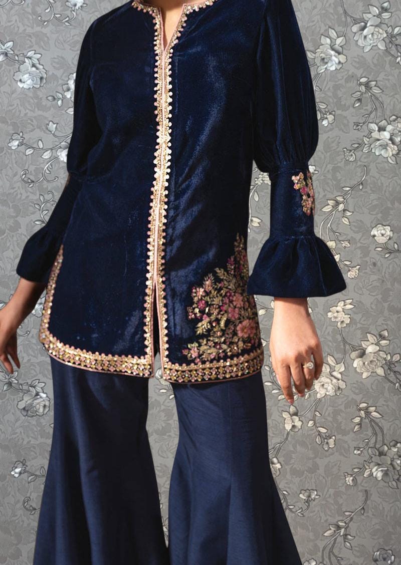 A woman wearing a navy blue velvet jacket with gold trim and floral embroidery on the sleeves, paired with matching blue pants.