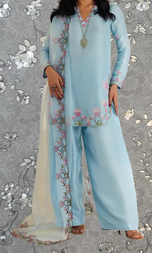 A woman wearing a light blue dress with floral embroidery and a matching shawl, standing against a gray floral background.