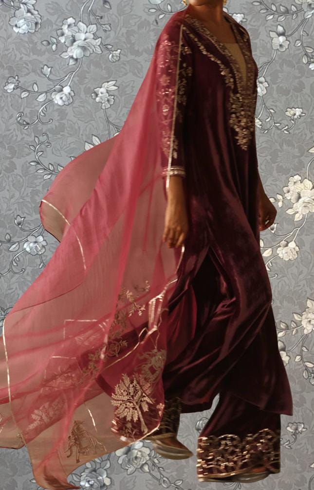 A woman wearing a long, burgundy velvet dress with a sheer pink veil, standing against a gray floral background.