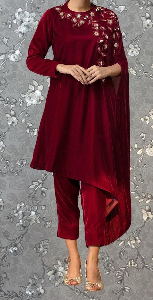 A woman wearing a red velvet dress with floral embroidery and gold heels stands against a gray floral background.