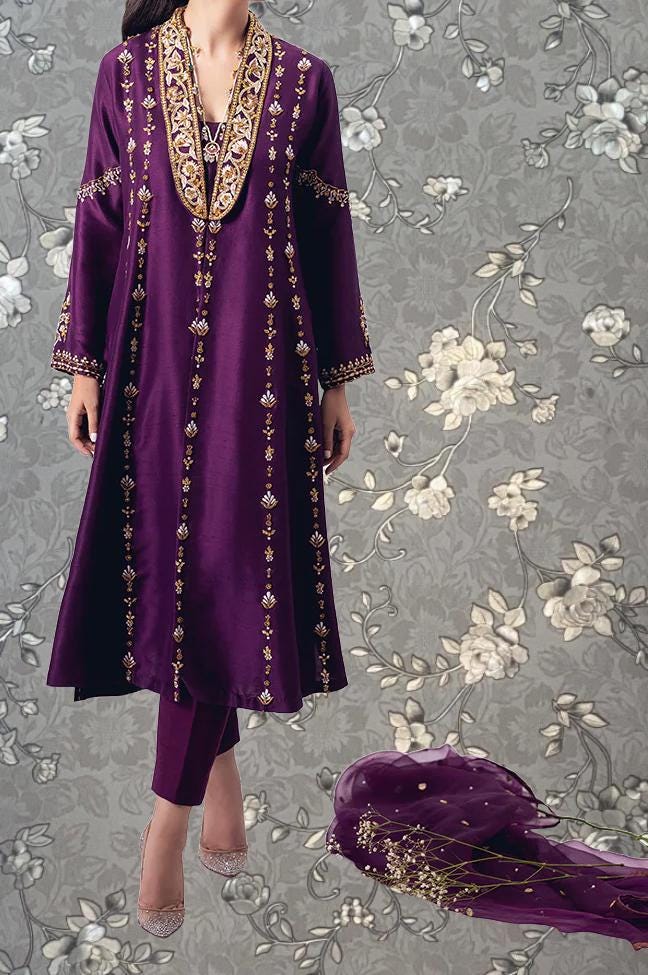 A woman wearing a long purple dress with gold embroidery stands against a gray floral background.
