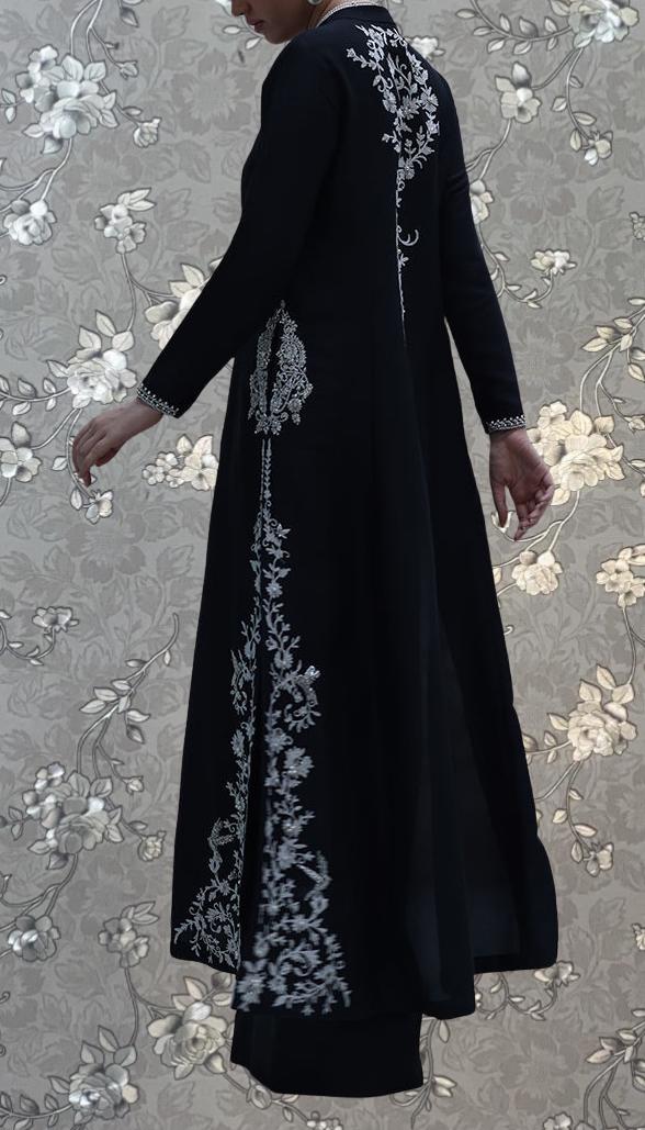 A person wearing a long black dress with intricate white embroidery stands against a floral patterned background.