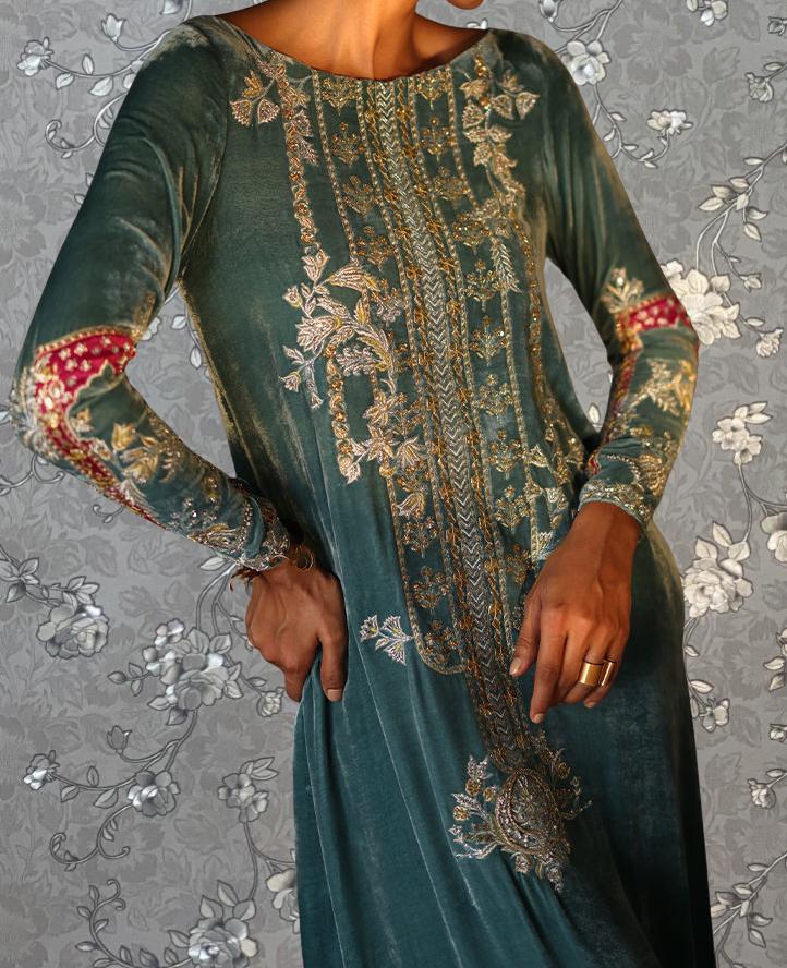 A woman wearing a long, green dress with intricate gold embroidery stands against a gray floral background.