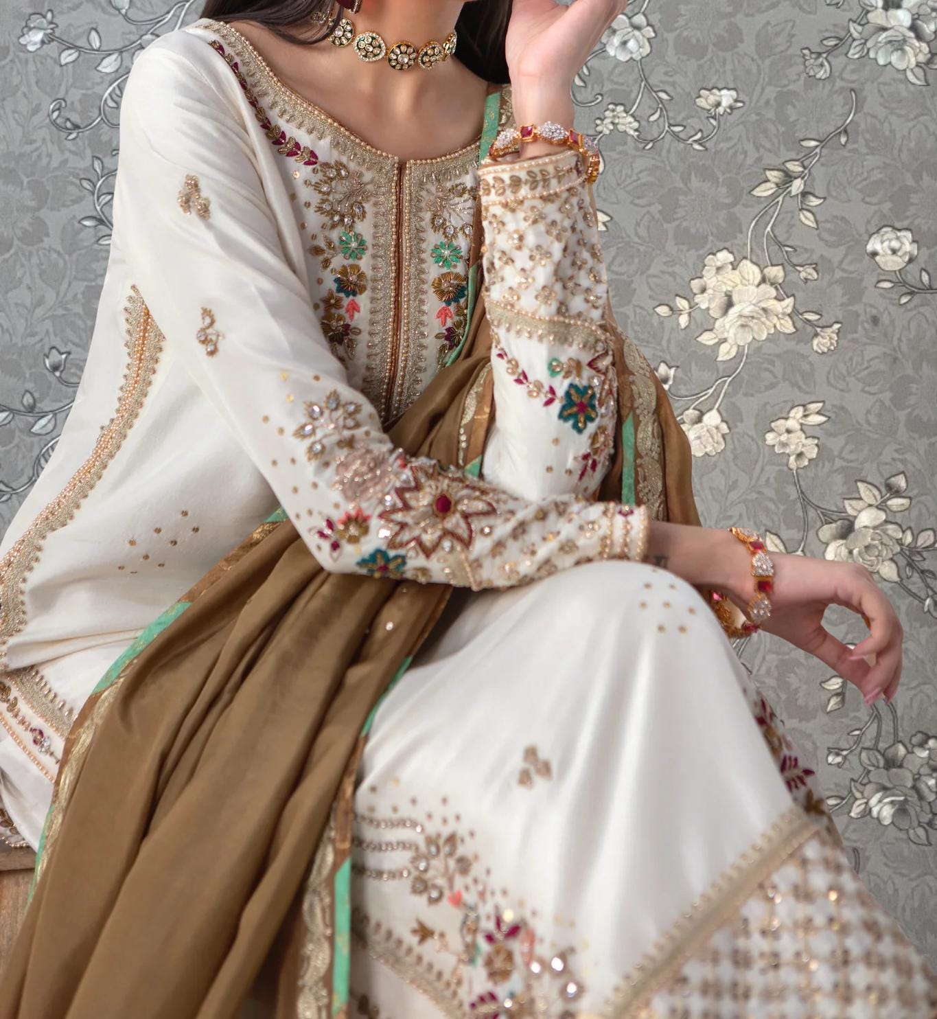 A woman wearing a white and gold dress with intricate embroidery and jewelry poses against a floral background.