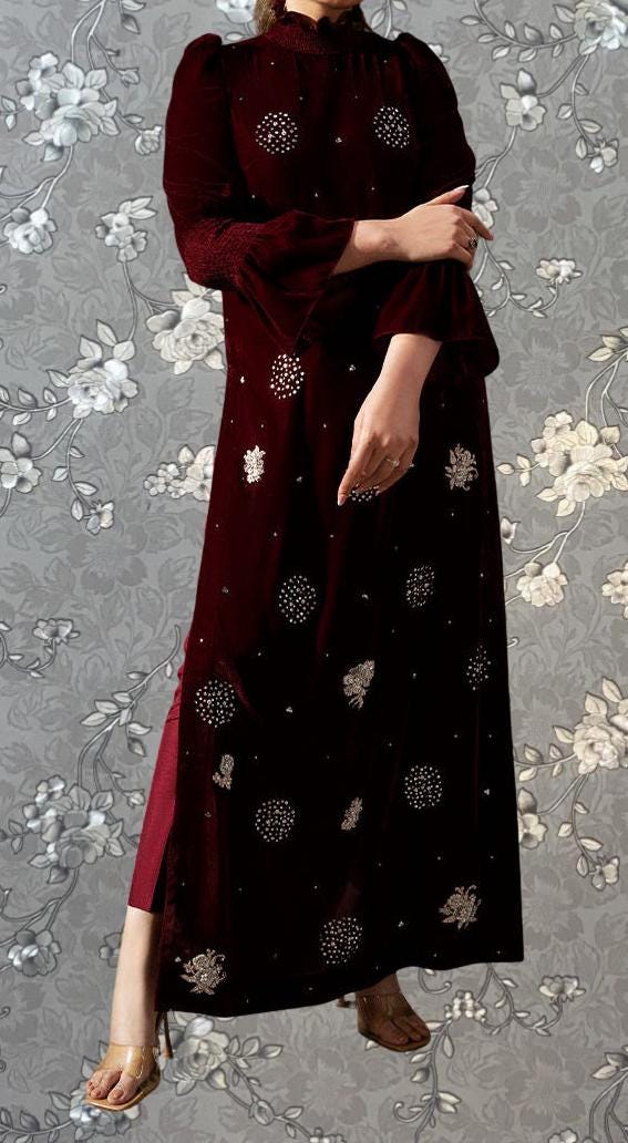 A person wearing a long burgundy dress with white floral embroidery stands against a gray floral background.