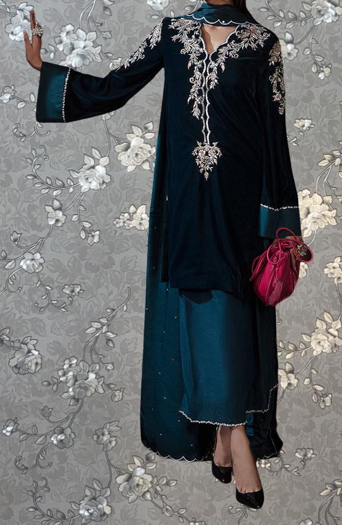 A woman wearing a long, dark blue dress with intricate embroidery and a matching purse stands against a gray floral background.