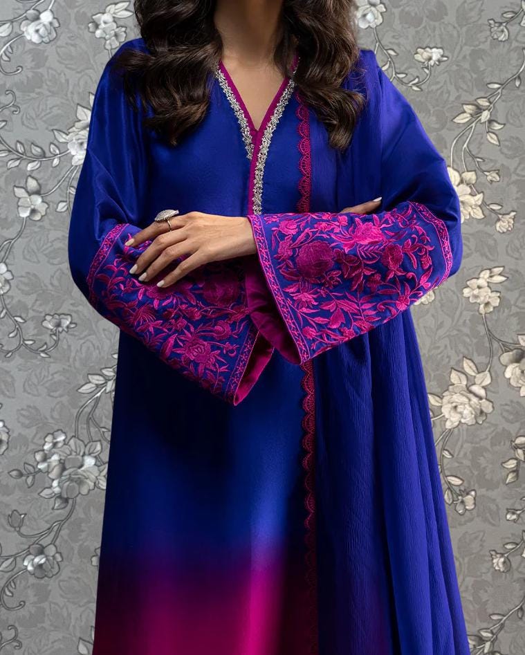 A woman wearing a long, blue dress with pink embroidered sleeves stands against a gray floral background.