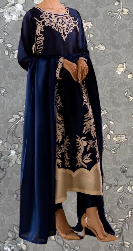 A woman wearing a navy blue dress with gold embroidered patterns, standing against a gray floral background.