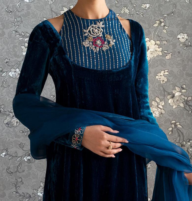 A woman wearing a blue velvet dress with a floral pattern and a high neckline, posing against a gray floral background.