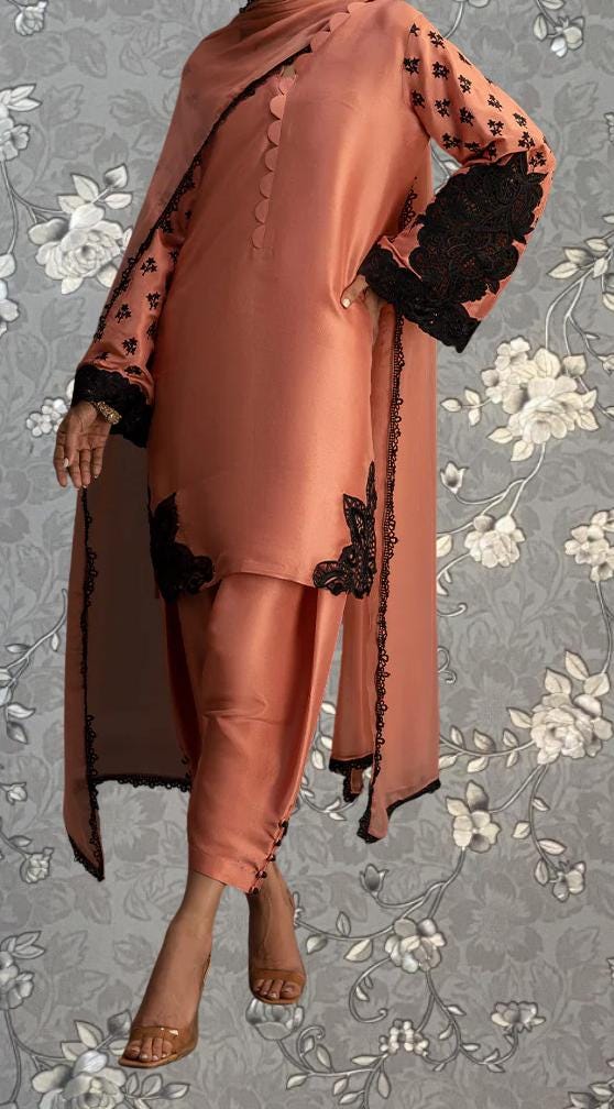 A woman wearing a peach-colored dress with black floral accents, standing against a gray floral background.