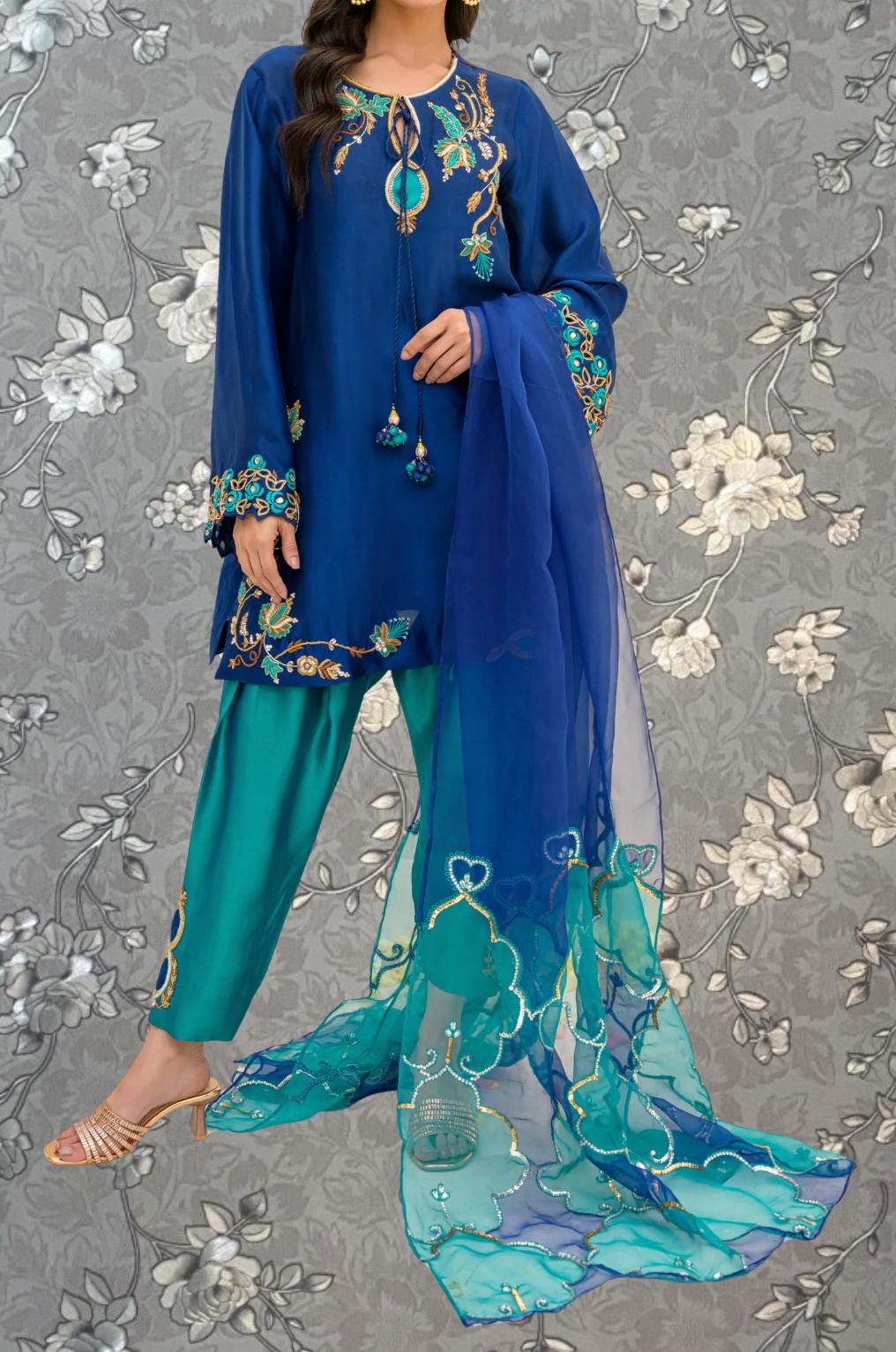 A woman wearing a blue dress with intricate embroidery and a matching blue shawl, standing against a gray floral background.