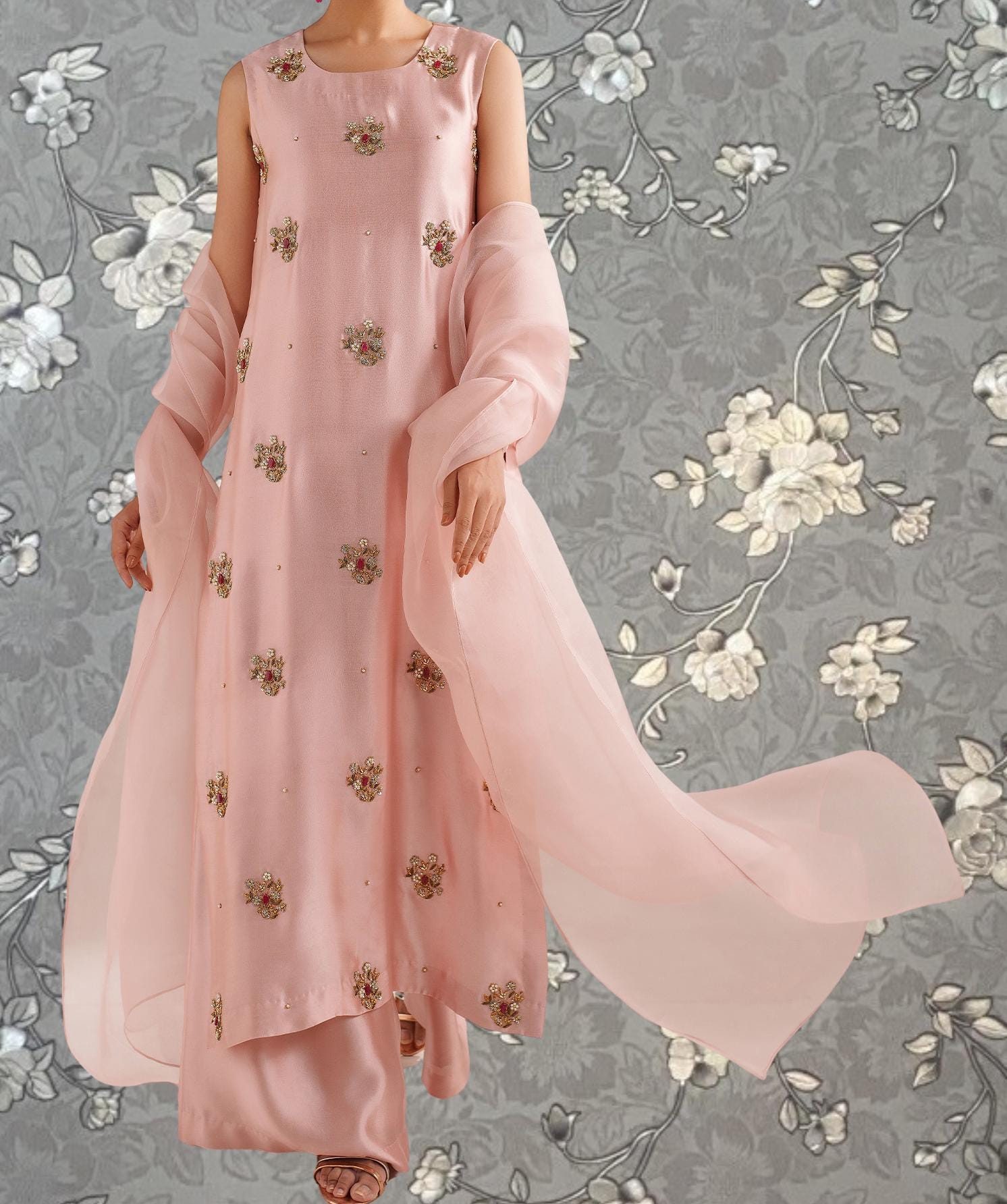 A woman wearing a pink dress with floral embroidery stands against a gray floral background.