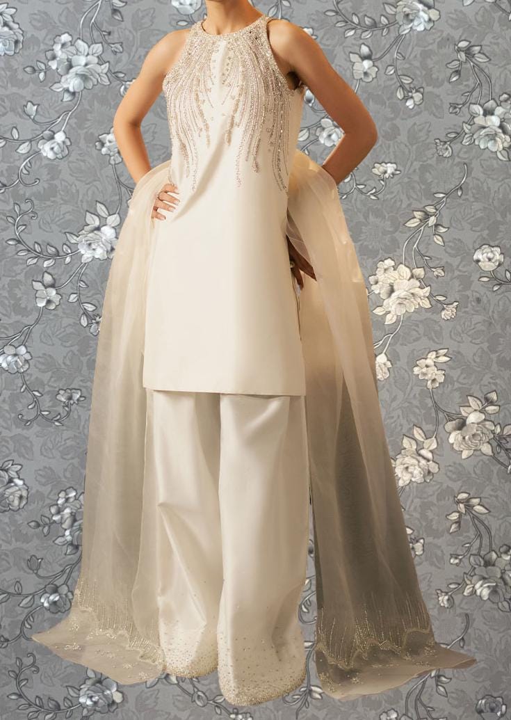 A woman wearing a white dress with a beaded bodice and a long, flowing skirt stands against a gray floral background.