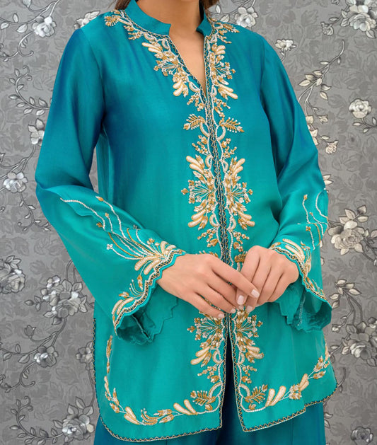 A woman wearing a teal-colored dress with intricate gold embroidery on the sleeves and collar.