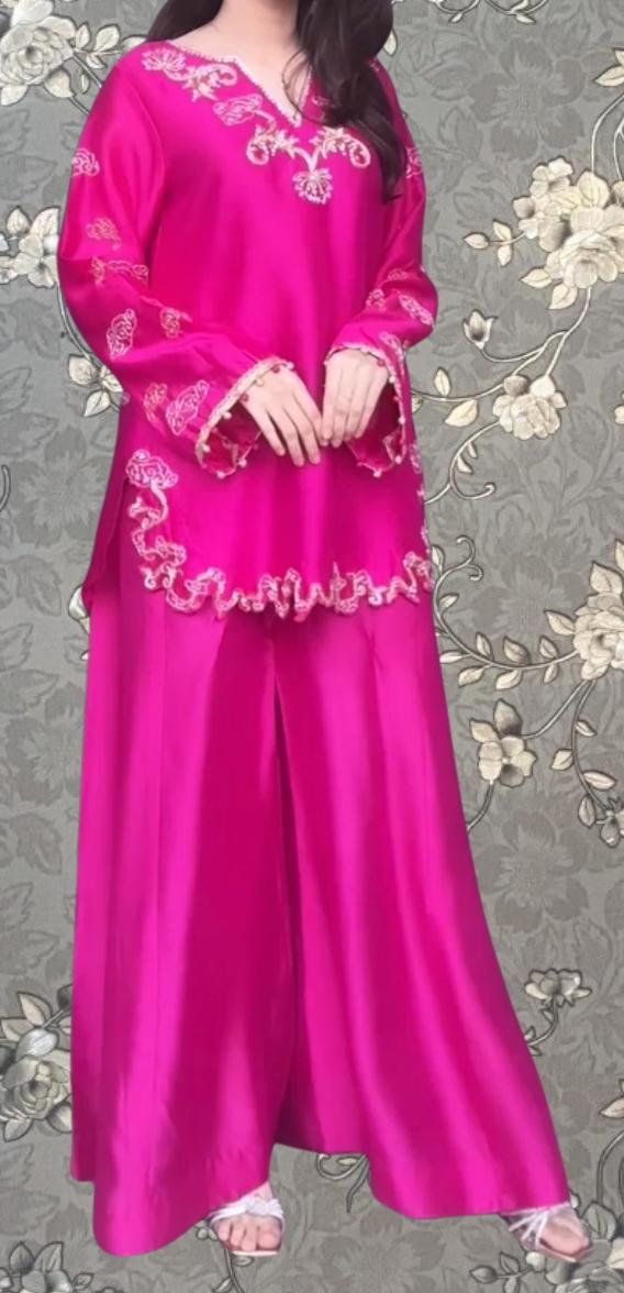 A woman wearing a long pink dress with intricate embroidery stands against a floral background.