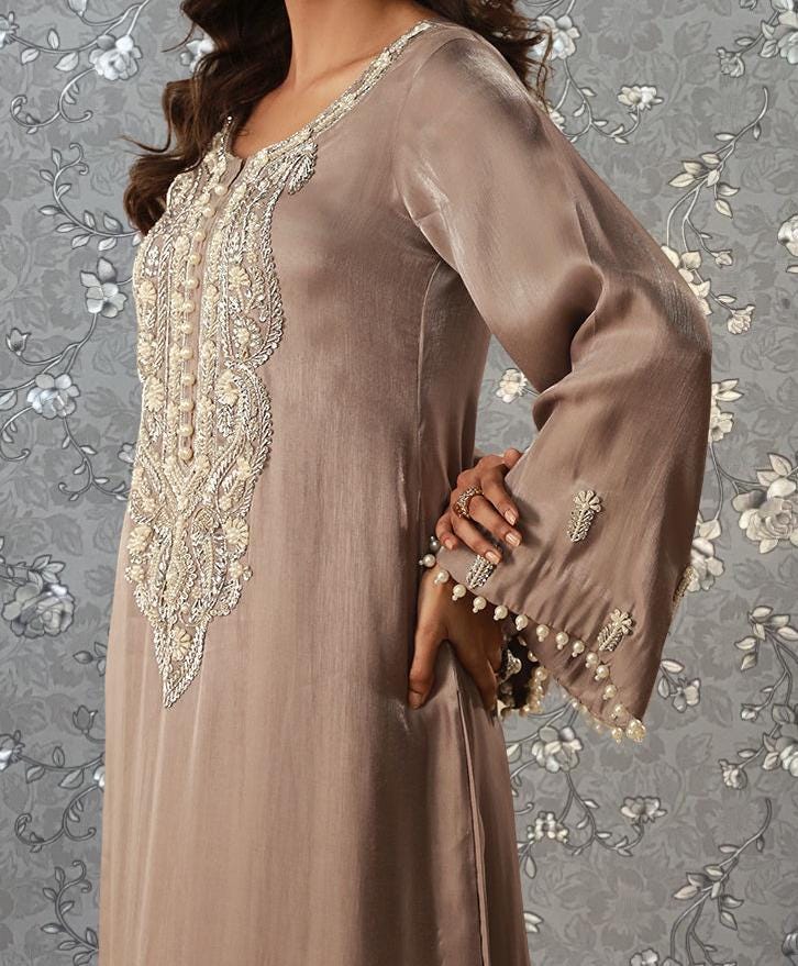 A woman wearing a long, beige dress with intricate embroidery and pearls on the sleeves.