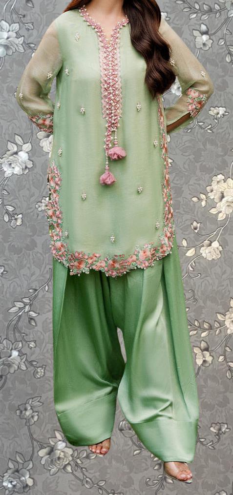 A woman wearing a green dress with pink accents and floral embroidery, standing against a gray floral background.
