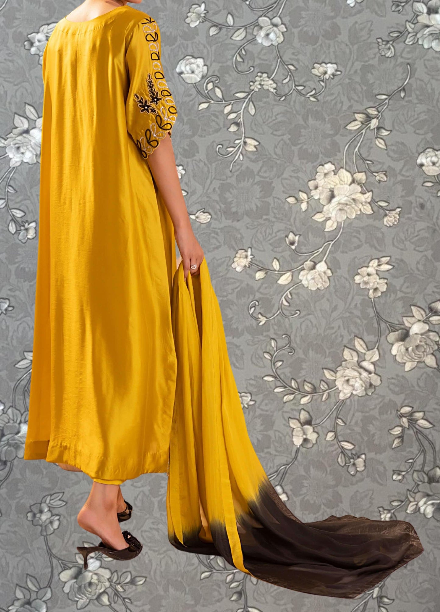 A person wearing a long, yellow dress with floral embroidery stands against a gray floral background.