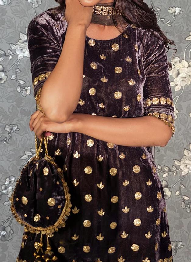 A woman wearing a purple velvet dress with gold polka dots and a choker necklace stands against a gray floral background.