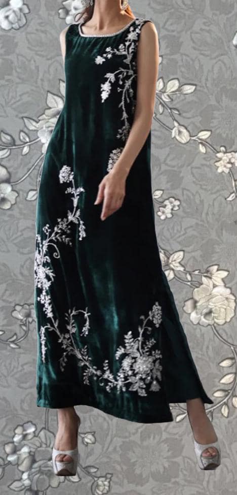 A woman wearing a long, sleeveless green dress with white floral embroidery stands against a gray floral background.