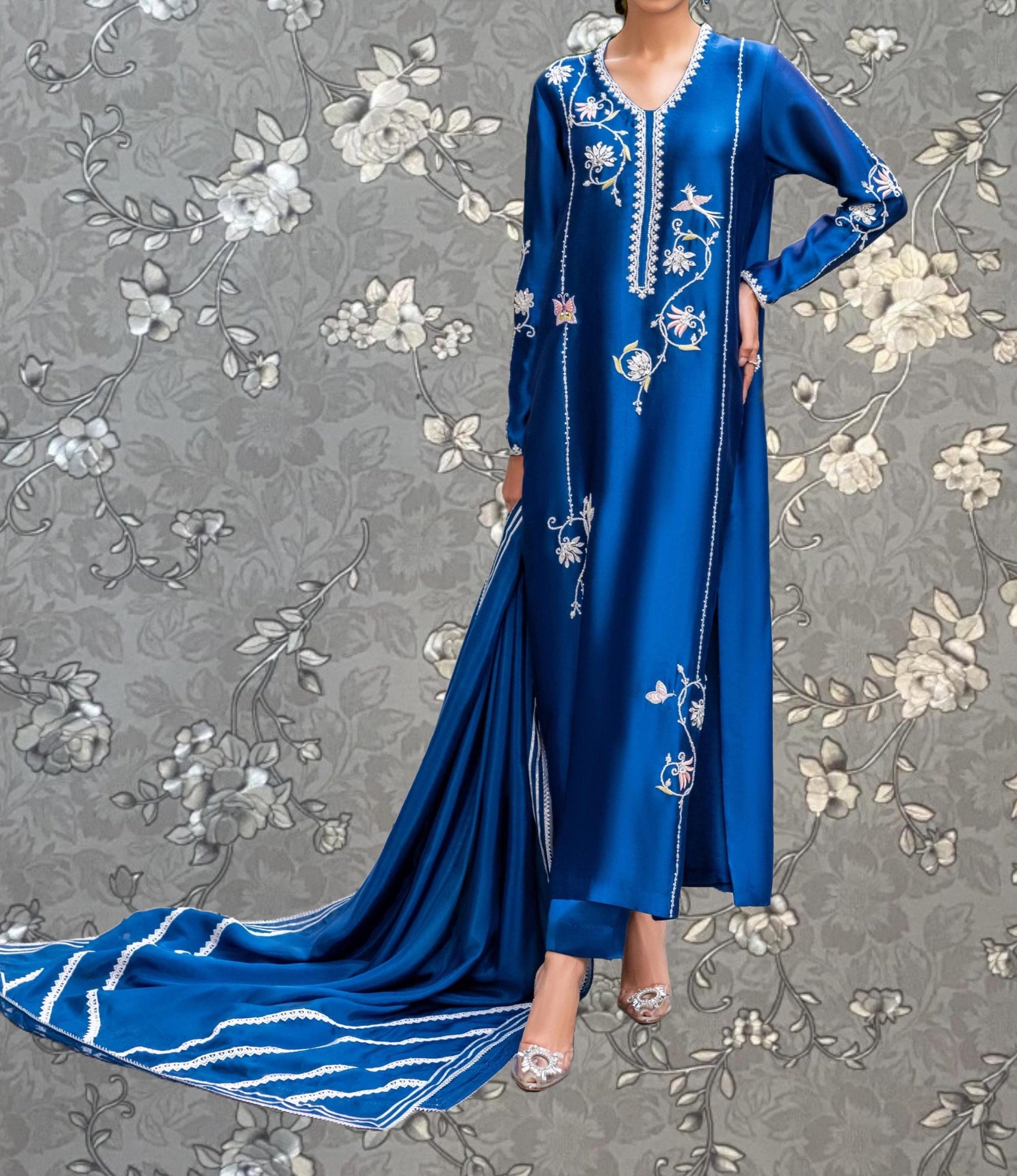 A woman wearing a long, blue dress with intricate embroidery stands against a gray floral background.