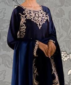 A woman wearing a blue dress with white embroidered patterns on the bodice and sleeves, standing against a gray floral background.