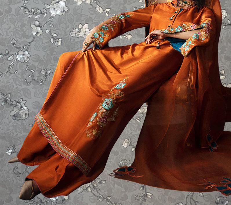 A woman wearing an orange dress with floral embroidery is sitting on a chair against a gray floral background.
