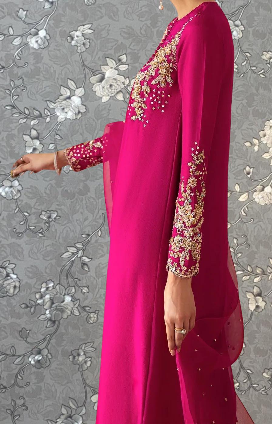 A woman wearing a long, pink dress with gold embroidery stands against a gray floral background.