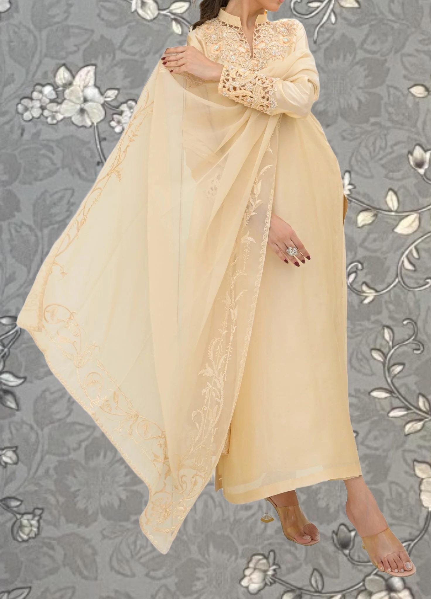 A woman wearing a long, cream-colored dress with intricate embroidery stands against a gray floral background.