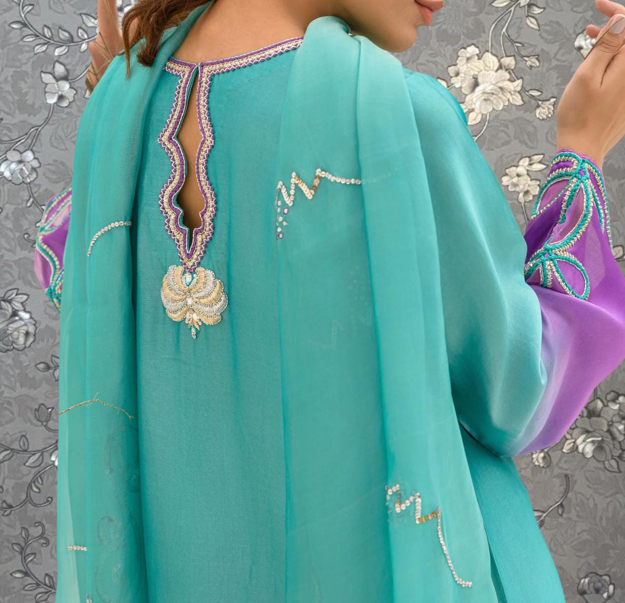 A close-up view of a woman wearing a turquoise dress with a purple and green patterned sleeve, posing against a floral background.