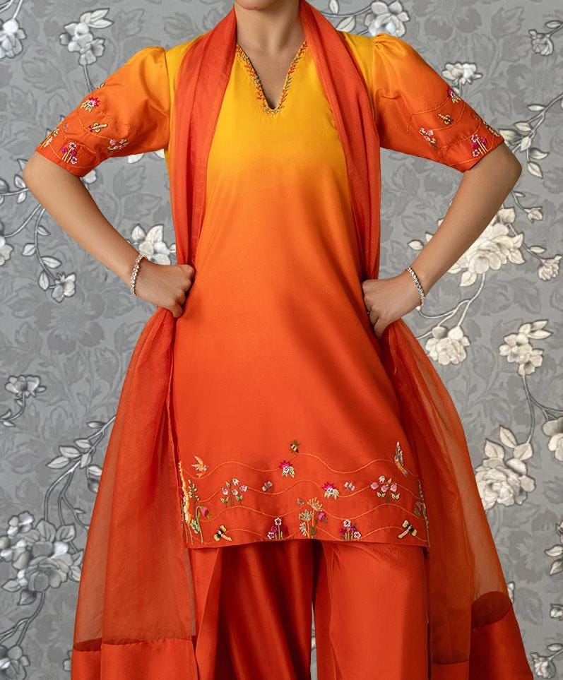A person wearing a vibrant orange and yellow dress with floral embroidery stands against a gray floral background.