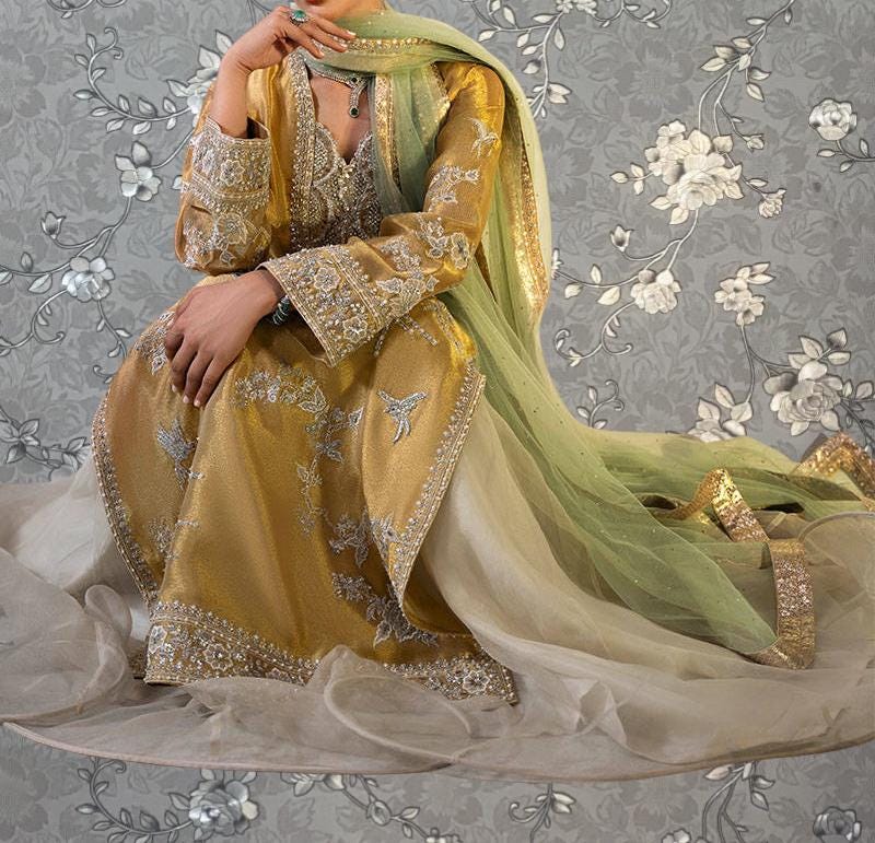 A woman wearing a gold and green traditional Indian outfit with intricate embroidery and a flowing green shawl, sitting on a white surface against a gray floral background.
