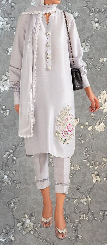 A person wearing a white dress with floral embroidery and a matching white scarf, standing against a gray floral background.