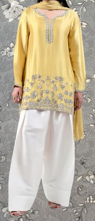 A person wearing a yellow dress with silver embroidery and white pants stands against a gray floral background.