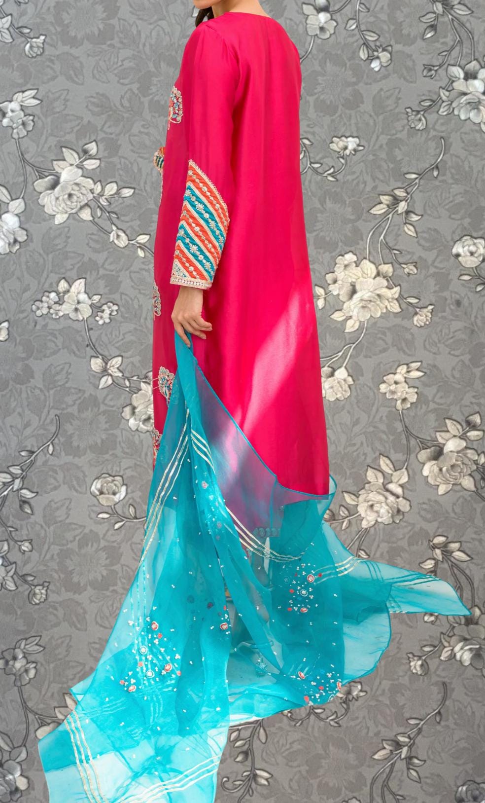 A person wearing a long pink dress with a turquoise shawl draped over their shoulders, standing against a gray floral background.