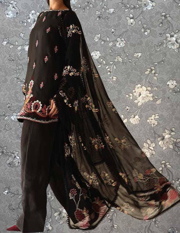 A person wearing a black dress with intricate floral embroidery stands against a gray floral background.