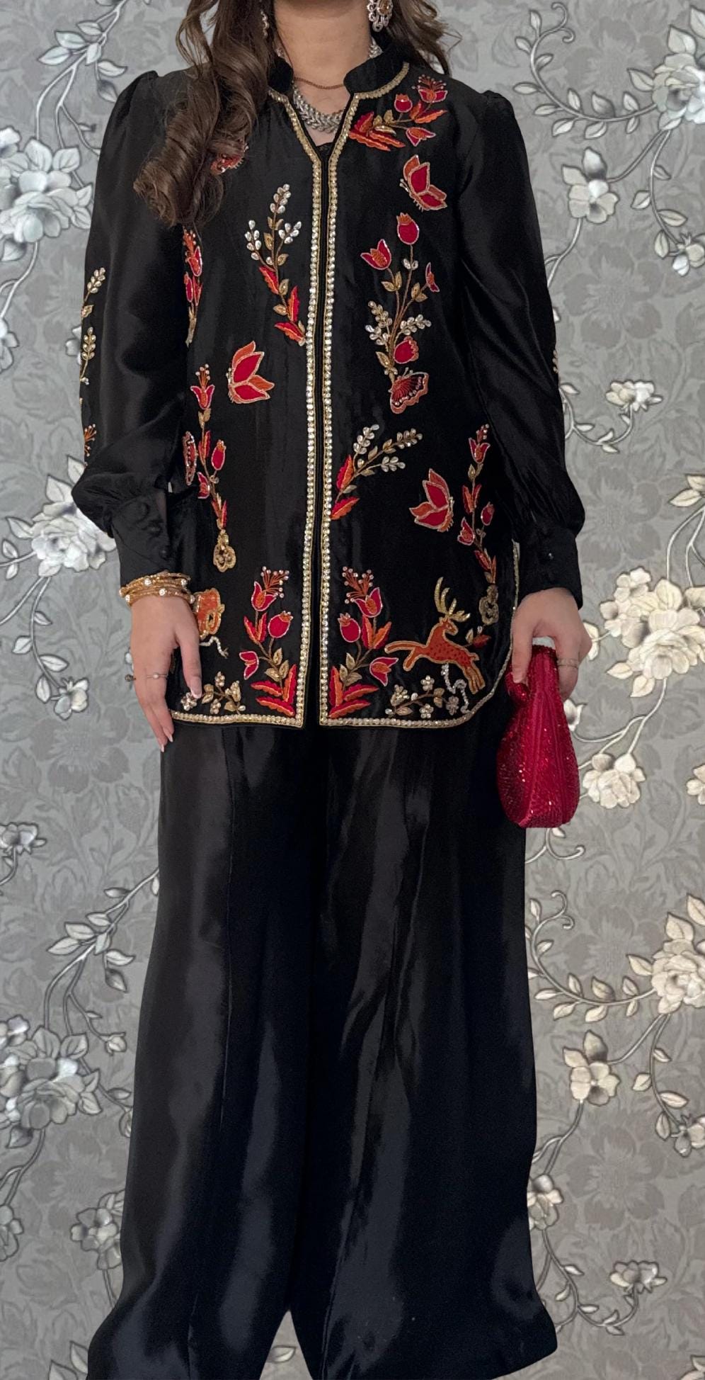 A woman wearing a black dress with red floral embroidery and holding a red clutch purse stands against a gray floral background.