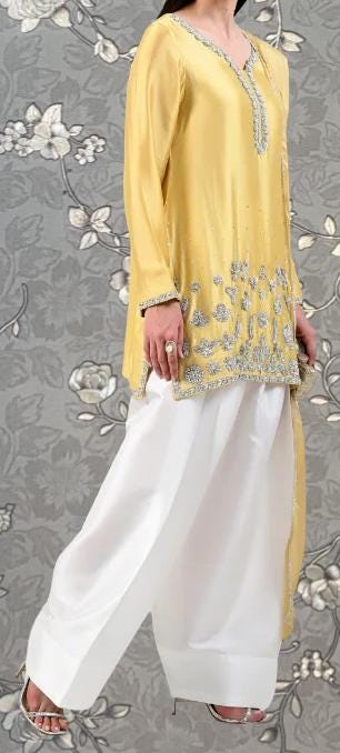 A woman wearing a yellow and white dress with intricate embroidery, standing against a gray floral background.