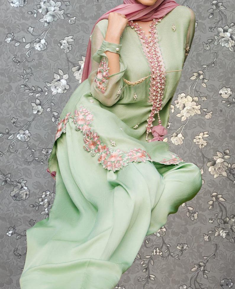 A woman wearing a green dress with pink floral embroidery and a pink scarf is shown against a gray floral background.