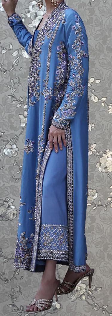 A woman wearing a long, blue dress with intricate embroidery and floral patterns, standing against a gray floral background.