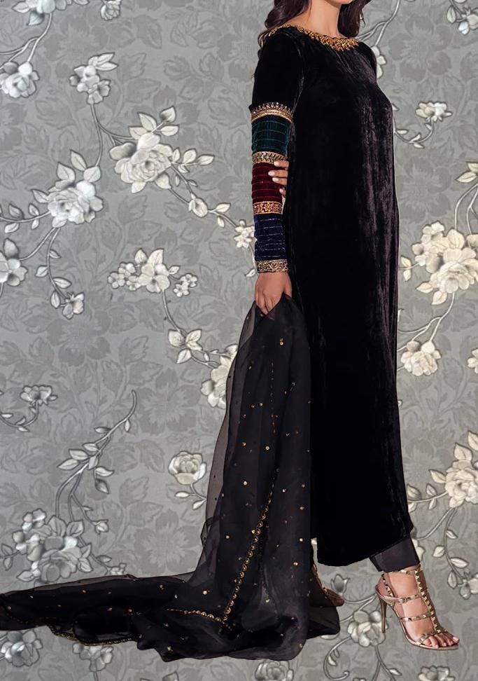 A woman wearing a long black velvet dress with gold accents and a matching black shawl stands against a gray floral background.