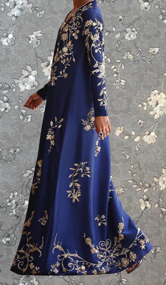 A woman wearing a long, blue dress with gold embroidery stands against a gray floral background.