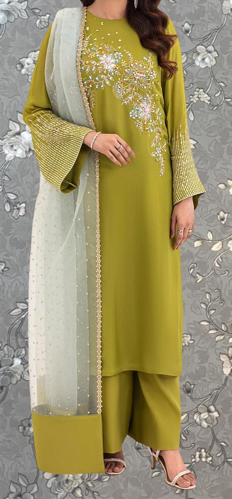 A woman wearing a green dress with gold embroidery and a matching green shawl stands against a gray floral background.