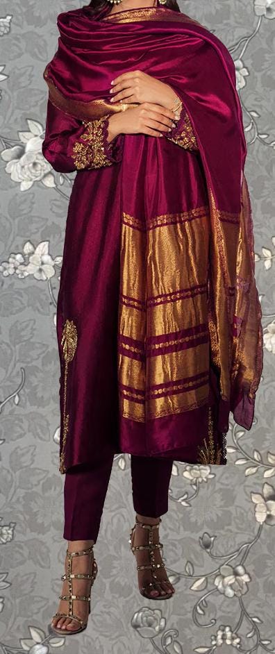 A person wearing a vibrant purple and gold outfit, including a long shawl and matching heels, stands against a gray floral background.