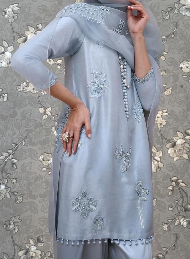 A person wearing a light blue dress with intricate embroidery and a matching light blue scarf stands against a floral background.