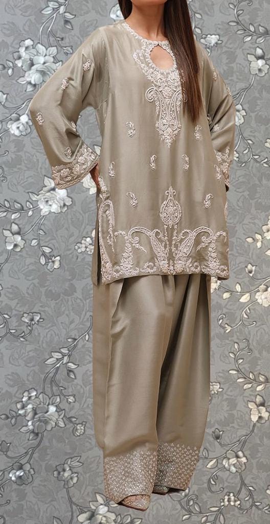 A woman wearing a beige dress with intricate embroidery and floral patterns, standing against a gray floral background.