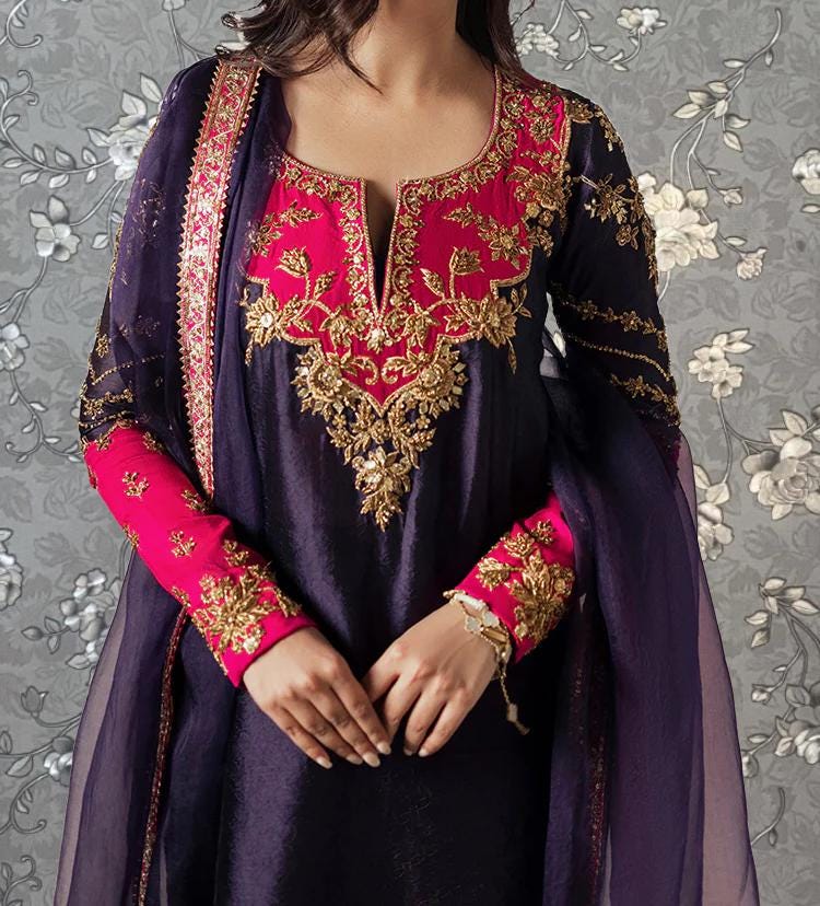 A woman wearing a purple and pink dress with intricate embroidery and gold accents, posing against a gray floral background.