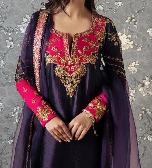A woman wearing a purple and pink dress with intricate embroidery and gold accents, posing against a gray floral background.