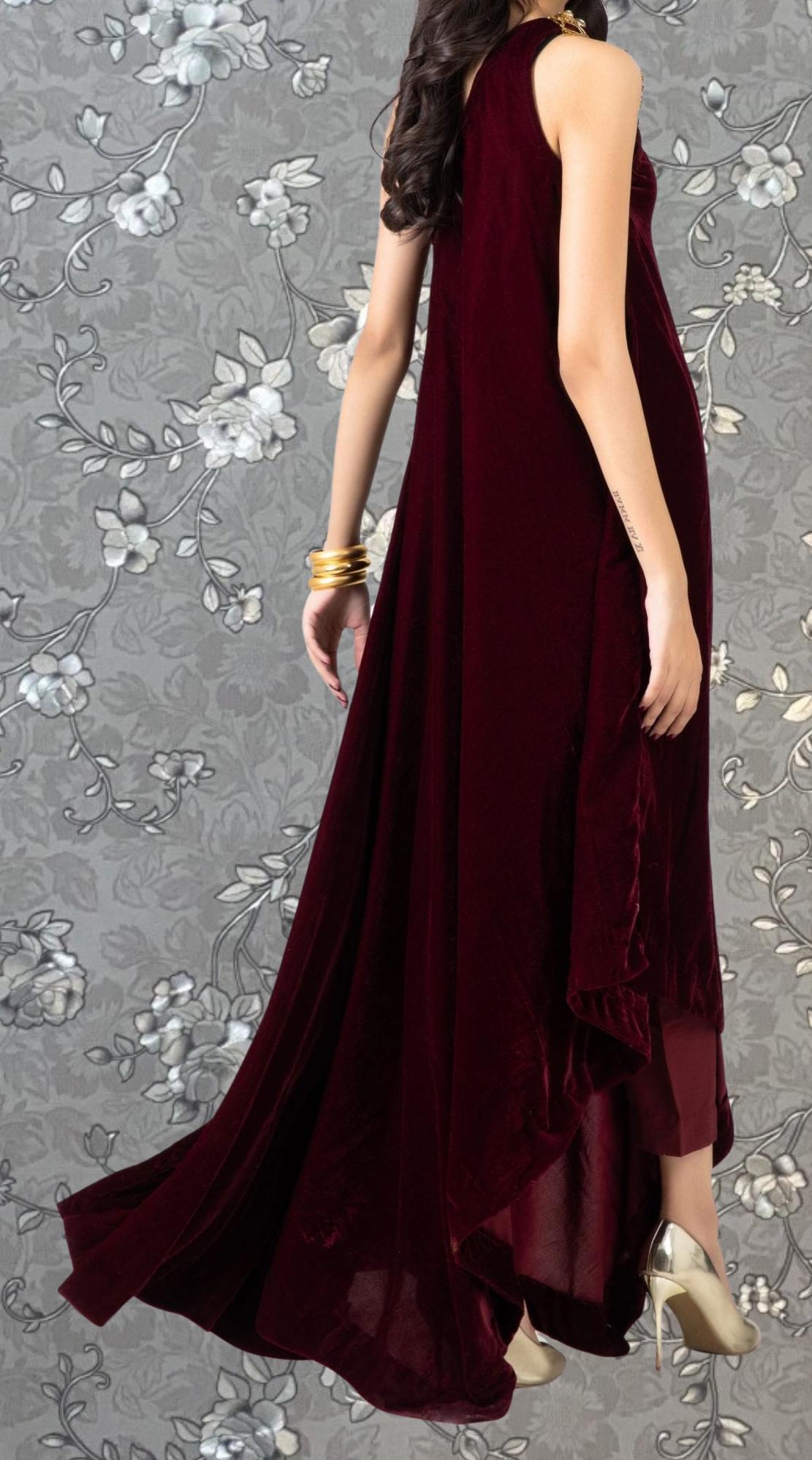 A woman wearing a long, burgundy velvet dress with a flowing train, standing against a gray floral background.