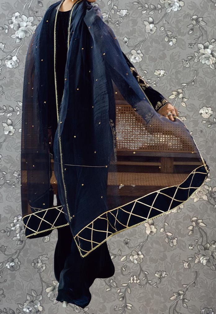 A person wearing a long, dark blue shawl with gold accents, standing against a gray floral background.